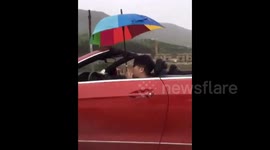 Man holds umbrella while driving convertible with the top down in the rain