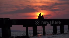 A man on a motorcycle on a bridge takes selfies with a stunning ocean sunset in the background.