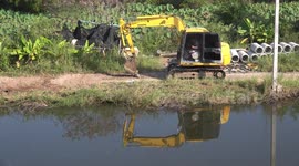 A mechanical digger working on a small country road by a lake, with reflections in water.