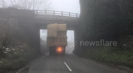 A tractor towing a  trailer load of big bale straw gets stuck under bridge