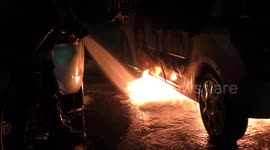 A car is burning and gasoline is dripping and burning. Firefighter sprays water and foam for extinguishing the fire