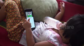 A kid with a milk bottle in one hand and a smart phone in the other falls asleep while playing games on her phone.