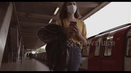 Young adult woman wearing protective medical face mask standing on underground train platform waiting and using mobile smart phone during coronavirus pandemic