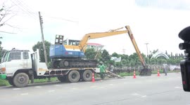 A large earth mover being unloaded from a tow truck, on  busy road in Thailand.