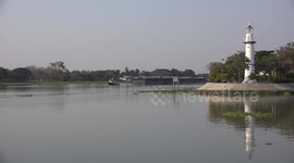 Boats and a barge sail past a white lighthouse on a river in Thailand.