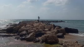 Girls sitting on a rocky beach, legs in the water, busy talking and looking at people kayaking with an active power plant running in the background. Fisherman's village Hadera