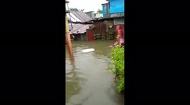 A woman carriying a kid jokingly greets the cameraman while walking through the flood water