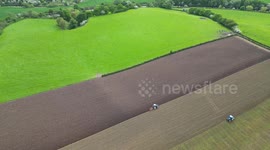 Drone shot of a dog running round multicoloured fields getting ploughed by two tractors facing opposite directions in the cheshire sun