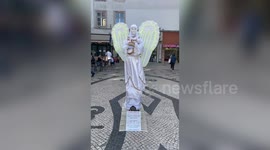 Creepy street performer dressed as angel holds moving baby doll