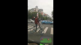 Chinese cyclist balances water bucket on head while riding one-legged across road