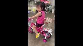 A 4-year-old girl rides a bicycle carrying a chicken on the back of her bicycle