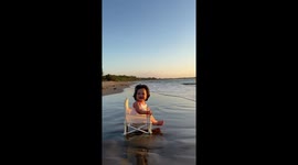 Cute baby enjoys waves on beach shore and sunset while sitting on small chair