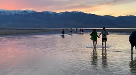 An unusual lake has formed in Death Valley's Badwater Basin (from extreme rainfall), and people have come to paddleboard and wade in this salty lake as the sun sets over the desert mountains, reflected in the waters of the lake.