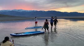 Paddleboarders head out as the sun sets on an unusual lake that has formed in Death Valley's Badwater Basin (from extreme rainfall). Tourists experience this salty lake as the sun sets over the desert mountains, reflected in the waters of the lake.