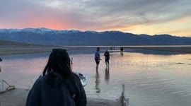 Paddleboarders head out, and visitors come from all over to see an unusual lake formed in Death Valley's Badwater Basin (from extreme rainfall. People paddleboard and wade in this salty lake as the sun sets over the desert mountains, reflected in the wate