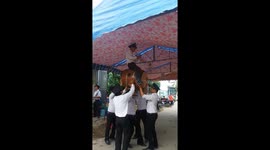 The man uses his head to lift a giant drum and another man above coordinates to blow the trumpet and play the drum