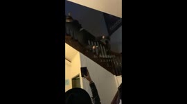 woman in black jumps over a 2nd floor wooden bannister and falls the the ground while her friends scream