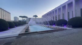 Purple lights at sunset illuminate the Piazzale delle Fontane and fountains spout water in front of the entrance to the Palazzo delle Fontane in the EUR district of Rome.