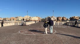 A man and a little girl pause and talk about the compass rose drawn with cobblestones on the pavement of the Pontile di Ostia while a man plays music on a clear December day in Rome.