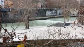 Winter landscape with bare branches around the Tiber flowing into the waterfall on Tiber Island and some people walking along the river bank on Christmas Day in Rome.