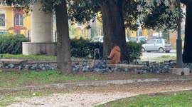 A woman sitting alone on a bench in a public park with a shopping trolley at her side surrounded by pigeons that finally take flight on the last day of the year in a suburb of Rome.
