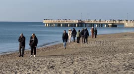 People walking on a stretch of beach at Lido di Ostia with calm sea, clear sky, in the background the pier of Ostia with people on the first day of the year in Rome.