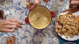 Group of friends eating fondue, a Swiss melted cheese dish served in a communal pot  over a portable stove heated with a candle and eaten by dipping bread into the cheese using long forks.
