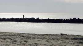 The silhouette of a man on the rocks fishing while a woman walks across the scene on the sand on the Fiumicino seafront near Rome.