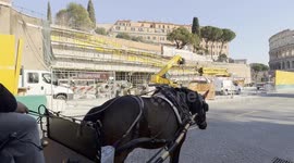 A botticella, a horse-drawn carriage, travels along via dei Fori Imperiali in front of the new Metro C stop under construction, with the Colosseum in the background and tourists walking along the street in Rome.