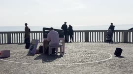 A man in a pink tracksuit plays the piano facing the sea with spectators leaning on the railing on the Lido di Ostia pier on a sunny February morning in Rome.