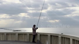 An elderly man is fishing on the Lido di Ostia pier on a February morning with calm seas and a grey sky that allows a few rays of sunshine to be glimpsed, in Rome.