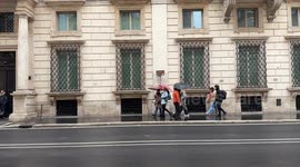 People with umbrellas walk along Via del Corso on a rainy winter day in Rome.