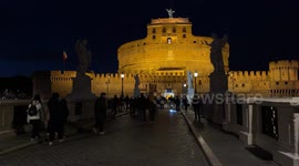 Tourists on Ponte Sant’Angelo with a few street lamps lit and Castel Sant’Angelo in the background on Christmas Eve in Rome.