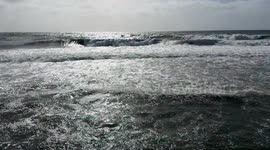 Wide shot of the motion of the waves of the very rough sea with reflections of the sun and cloudy sky on the horizon on the coast of Lido di Ostia in Rome.