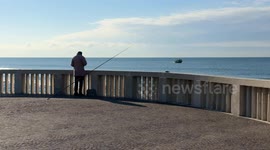 An elderly gentleman fishing on the pier of the Lido di Ostia adjusts his fishing line as a small fishing boat sails close to the coast of the Lido di Ostia on a morning with few clouds, blue sky and calm sea in Rome.