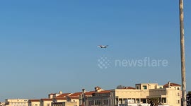 An American Airlines plane flies through the blue sky as it prepares to land at Fiumicino Airport and disappears behind some buildings in Rome.
