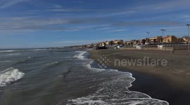 Wide shot of the waves on the coast as a man looks out to sea and his son plays with a ball on the beach of Lido di Ostia on a clear day in March in Rome.