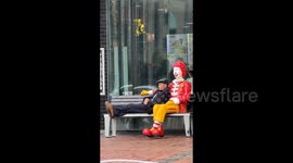 Elderly man casually rests in Ronald McDonald's arms on bench