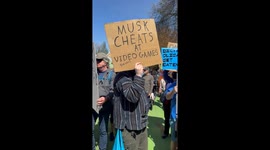 A protester at an anti-Trump anti-Elon Musk protest with a strange sign stating that Elon Musk cheats on video games
