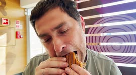A man is taking a big bite of a grilled burger inside a fast-food restaurant. He enjoys a casual meal in a relaxed atmosphere.
