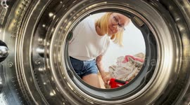 Point of view from inside a washing machine as a young woman in casual clothes loads laundry. Modern home scene with clear view through washer door