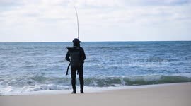 Solitary fisherman casting line into ocean from shore on a calm cloudy day, peaceful coastal fishing scene with waves and distant horizon.