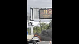 Close-up view of a burning car on the M1 near Milton Keynes, flames visible with thick smoke affecting all four lanes.