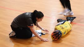 A young inline speed skating competitor lies on the ground after a fall during a tournament. A concerned coach or medic kneels beside, checking on the injured child inside a sports hall.