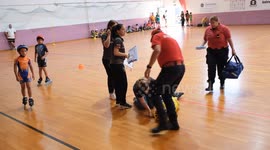 A young inline speed skating competitor lies on the ground after a fall during a tournament. A concerned coach or medic kneels beside, checking on the injured child inside a sports hall.