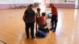A young inline speed skating competitor lies on the ground after a fall during a tournament. A concerned coach or medic kneels beside, checking on the injured child inside a sports hall.
