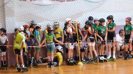 Young inline speed skaters wait behind the tape before the start of a children indoor tournament. Some sit on the floor, while others stand in colorful team uniforms, ready for the race.
