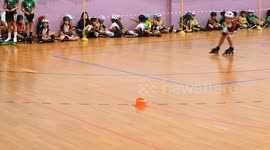 A young girl competes in an indoor inline speed skating tournament while other participants sit along the wall waiting for their turn. Bright lighting and floor markings define the track area.
