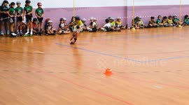 A young girl competes in an indoor inline speed skating tournament while other participants sit along the wall waiting for their turn. Bright lighting and floor markings define the track area.