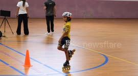 A young boy glides on one foot during an indoor inline skating event, confidently balancing and enjoying the ride. The scene takes place on a marked hardwood floor near cones and timers.
